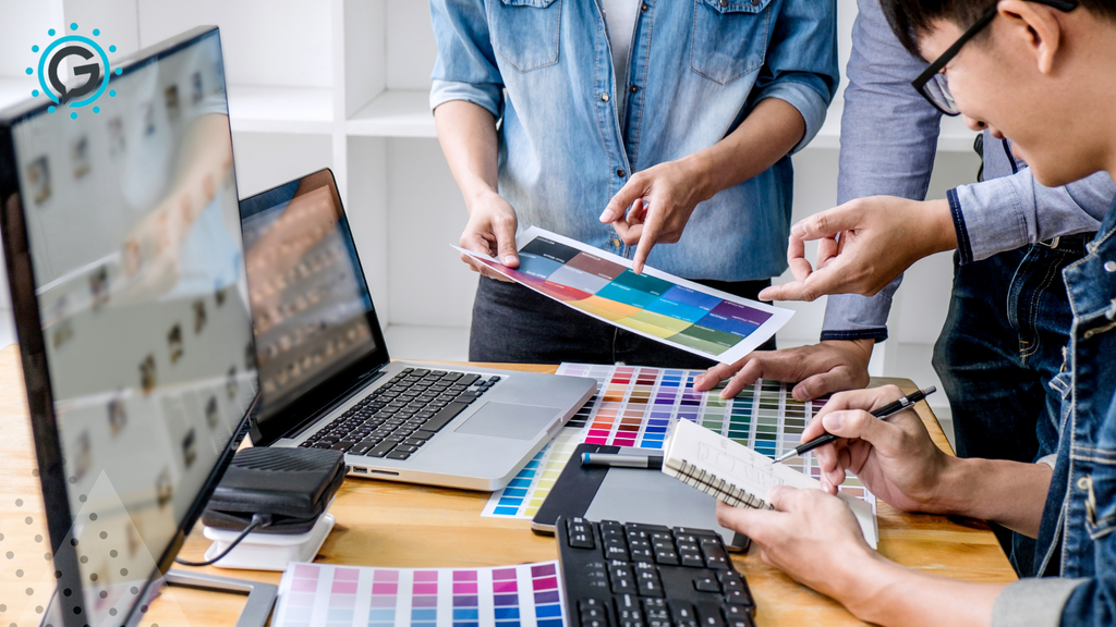 Three people stand around a desk looking at color swatches on paper. On the desk is also a laptop, monitor, and keyboard.
