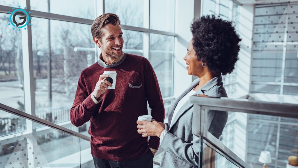 A white man in a red sweater smiles and hold a cup of coffee while speaking to a black woman with a short afro holding a cup of coffee wearing a grey blazer. They stand on a flight of stairs in front of large windows.