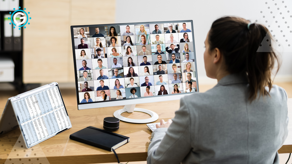 A white women with a brown ponytail in a grey blazer sits at a light wood desk looking at her computer monitor with about 64 faces on the call, each in their own square. On the desk there is a calendar and a black notebook.
