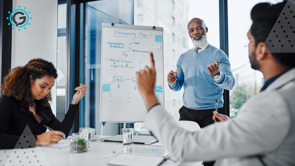 One dark haired man lifts his finger in the air to ask a question while his dark curly-haired coworkers sits opposite him at a white desk taking notes. An older black man with a white beard stands in front of them presenting something on a whiteboard.