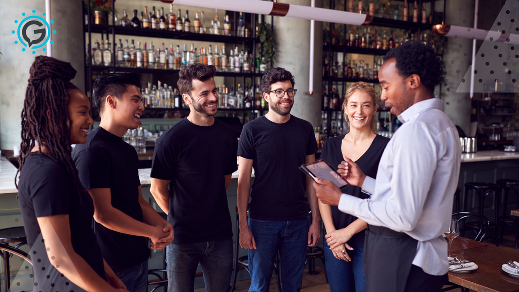 Two women and three men all wearing black t-shirts and jeans smile around a man in a white button up and apron reading off of a tablet.