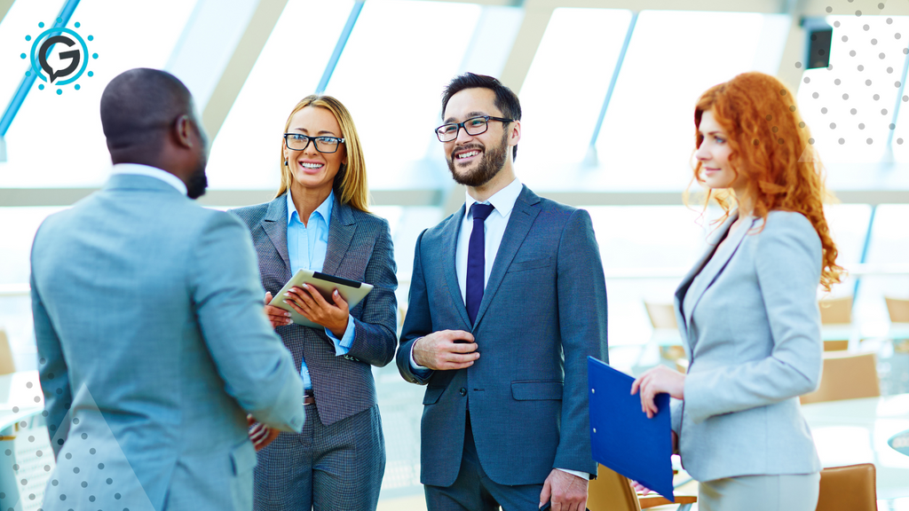 One black man in a grey suit talks to two white women and one white man, all in grey suits and smiling.