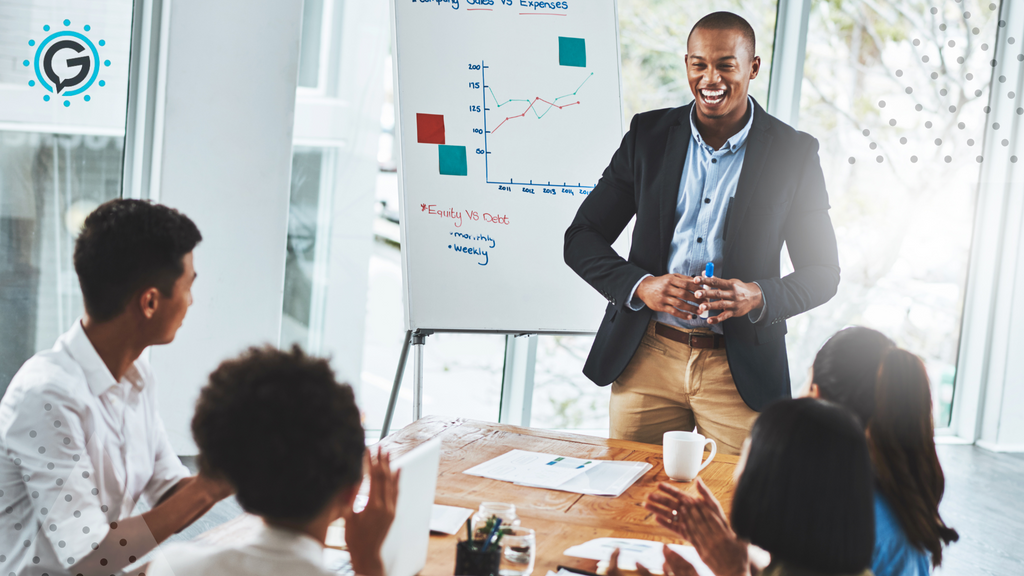 A black man in a navy blazer, light blue button up, and khaki pants smiling in front of a poster board with a graph on it, standing in front of four coworkers sitting at a desk and clapping.