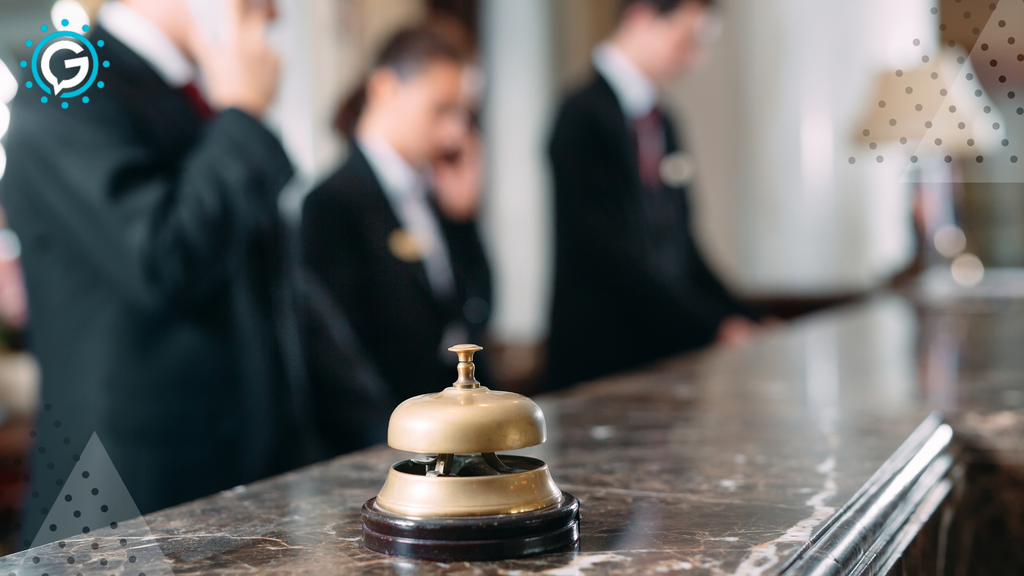 A gold desk call bell sits on a granite surface with three employees in the background talking on phones and wearing black blazers.