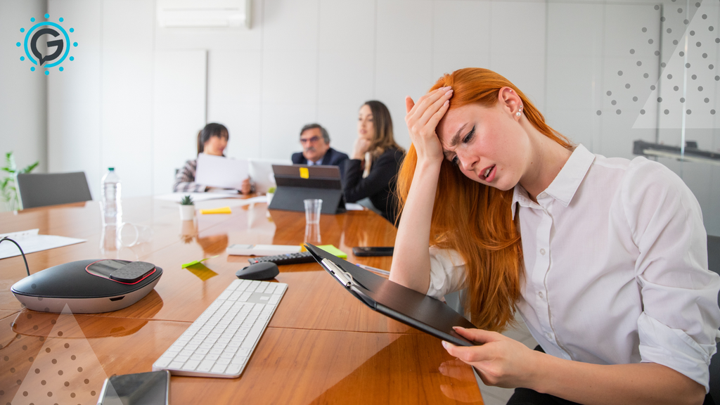 A white woman with red hair looks stressed with her head in her hand looking at a clipboard while sitting at a wood conference table as three coworkers talk behind her.