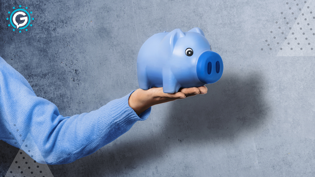 A blue piggy bank held in the hand of someone wearing a blue sweater. The piggy bank and the person holding it are in front of a faded grey backdrop.