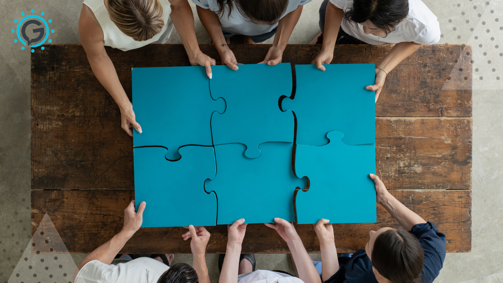 From bird's eye view, 6 people sitting around a desk, each holding a large blue puzzle piece and connecting it with the others, together creating a blue rectangle.